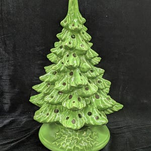 May include: A ceramic Christmas tree in a vibrant green color. The tree has a classic design with multiple tiers of branches and small holes for lights. It sits on a matching green base, creating a festive holiday decoration.