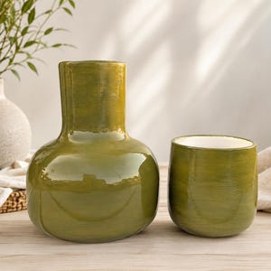 May include: A glossy olive green ceramic vase with a wide base and narrow neck, paired with a matching cylindrical olive green ceramic cup featuring a white interior. Both items have a smooth, hand-painted finish with visible brush strokes.