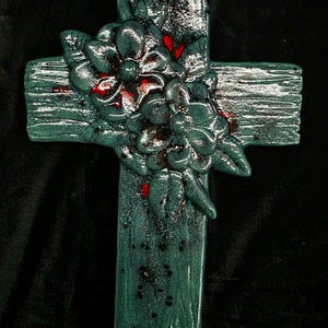 May include: A teal-colored cross with a textured, wood-like appearance. The cross is adorned with a cluster of sculpted flowers and leaves, accented with red details. The overall design has a rustic, handcrafted aesthetic.