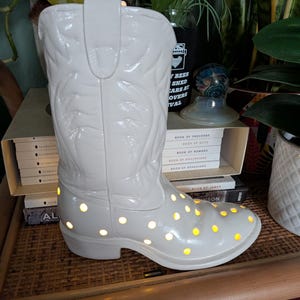 May include: A white ceramic cowboy boot-shaped lamp with decorative embossed details. The boot has numerous yellow lights illuminating from within. The lamp is a decorative item, suitable for home decor.