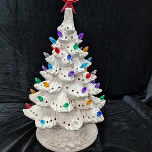 May include: A white ceramic Christmas tree with colorful lights and a red star on top. The tree has a round base with a holly leaf design. The lights are various colors, including red, blue, green, and purple.