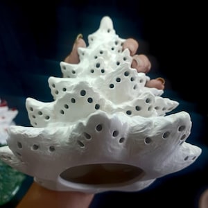 May include: A white ceramic Christmas tree with a textured surface and numerous circular cutouts. The tree has a tiered design, with each layer decreasing in size towards the top. The tree is held by a person.