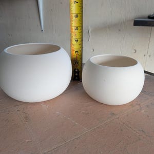 May include: Two white, spherical ceramic bowls of different sizes. The larger bowl is approximately 6 inches in diameter. The bowls have a smooth, matte finish and a neutral interior. They are suitable for various uses, such as planters or decorative items.
