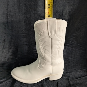May include: A white cowboy boot, approximately 10 inches tall, with decorative stitching. The boot has a classic western design, featuring a pointed toe and a stacked heel. The boot is made of a smooth, solid white material.
