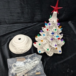 May include: A white ceramic Christmas tree with colorful lights and a red star topper. The tree is accompanied by a base and a bag containing a power cord and light bulb. The tree is a festive holiday decoration.