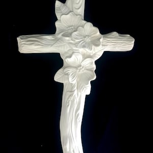 May include: A white, decorative cross with a floral design. The cross is made of a light-colored material and features detailed carvings of flowers and leaves. The background is solid black.