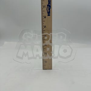 May include: A clear, plastic Super Mario logo with a wooden ruler measuring its height. The logo is approximately 4 inches tall. The ruler is marked with black lines and numbers, and the logo is transparent.