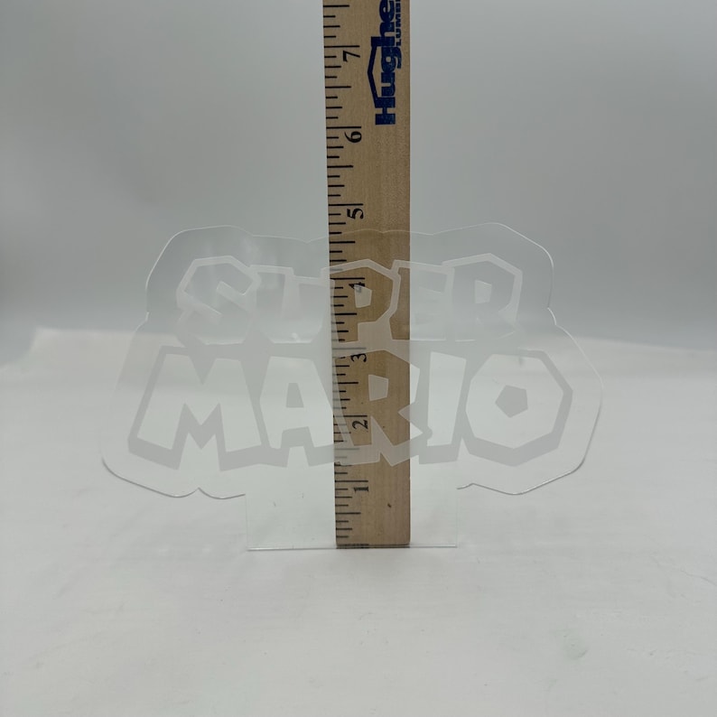 May include: A clear, plastic Super Mario logo with a wooden ruler measuring its height. The logo is approximately 4 inches tall. The ruler is marked with black lines and numbers, and the logo is transparent.