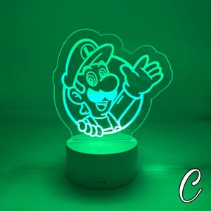 May include: A green-glowing acrylic LED lamp featuring a cartoon image of Luigi from the Super Mario Bros. video game series. The lamp has a white base and a detailed outline of Luigi's face, hat, and hand. The background is a solid green.