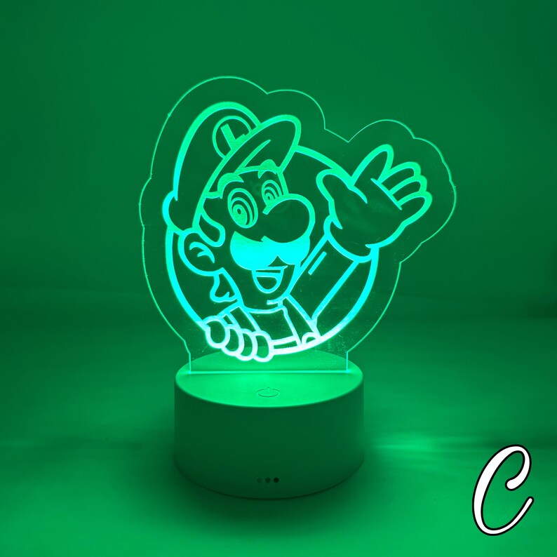May include: A green-glowing acrylic LED lamp featuring a cartoon image of Luigi from the Super Mario Bros. video game series. The lamp has a white base and a detailed outline of Luigi's face, hat, and hand. The background is a solid green.