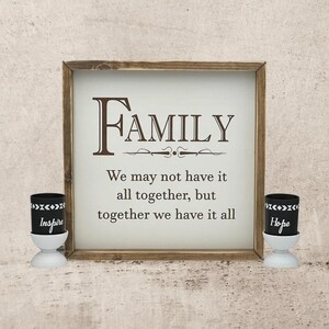 May include: A wooden sign with the word "FAMILY" in large letters and the quote "We may not have it all together, but together we have it all."  Two small black candle holders with the words "Inspire" and "Hope" are on either side of the sign.