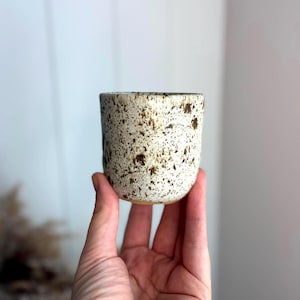 May include: A small, cylindrical ceramic cup featuring a cream-coloured base with a dense pattern of dark brown speckles throughout the exterior surface. The cup has a smooth, rounded rim and a straight-sided profile without a handle.