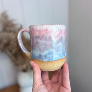 May include: A ceramic mug featuring a gradient glaze that transitions from a soft pink at the top to a light purple in the middle and a pale blue at the base, ending in a raw, unglazed tan clay bottom with a curved handle on the left side.