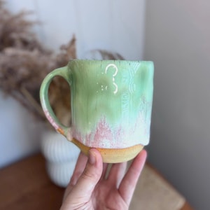 May include: A ceramic mug with a rounded handle, featuring a glossy glaze that transitions from a pale mint green at the top to a soft pink hue near the base, which remains an unglazed, textured tan clay colour at the very bottom edge.