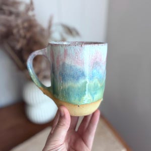 May include: A ceramic mug featuring a reactive glaze with a colour gradient transitioning from a pale pink at the rim to a deep blue in the centre and a vibrant green near the base, which is finished in a natural, unglazed tan clay texture.