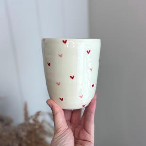 May include: A cream-coloured ceramic tumbler featuring a scattered pattern of small, hand-painted hearts in shades of bright red and soft pink. The vessel has a smooth, cylindrical shape with a slightly tapered base and a clean, open top rim.