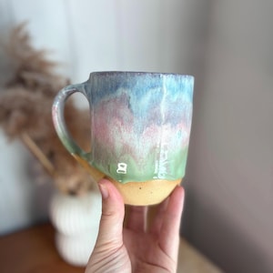May include: A ceramic mug featuring a reactive glaze with blended layers of blue, pink, and green colours. The upper portion of the mug displays a smooth, glossy finish, while the base remains unglazed with a natural, textured tan clay surface.
