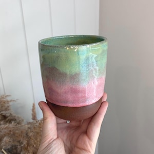 May include: A cylindrical ceramic cup featuring a smooth, unglazed dark brown base that transitions into a vibrant, glossy gradient of pink, soft purple, and light green hues across the upper body and the interior rim of the vessel.