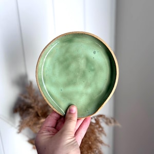 May include: A round ceramic spoon rest with a smooth, light green glazed interior surface and a speckled texture. The outer rim and base of the dish feature an exposed, unglazed tan clay finish with a natural, earthy appearance and rounded edges.