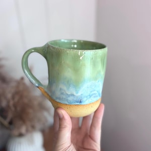 May include: A ceramic mug featuring a smooth, glossy glaze that transitions from a light green at the top to a vibrant blue in the middle, ending in a textured, unglazed tan base. The mug has a curved handle and a rounded, organic shape.