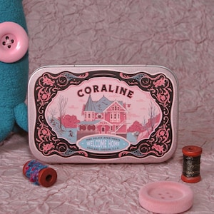 Puede incluir: Una lata metálica rectangular con la ilustración de una casa rosa y el texto Coraline y Pink Palace Apartments Welcome Home Ashland, Oregon, U.S.A. La lata está rodeada por un peluche azul, un carrete de madera y un botón rosa.
