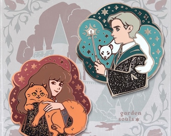 May include: Two enamel pins featuring characters from a fantasy series. One pin shows a person holding an orange cat, set against a red and brown background. The other pin depicts a person with a wand and a white ferret, set against a teal background.