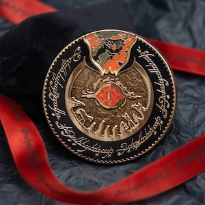 May include: A round, gold-colored metal coin with a black border and intricate design. The coin features a central red eye, surrounded by detailed artwork and script. A red ribbon and black velvet pouch are in the background.
