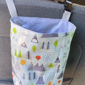 Hanging bag with buckle strap for organizing car or holding litter. Fabric is waterproof oilcloth. Pattern is blue and white interior with a    outside pattern of mountains and trees. Colors are green black and orange. Car seatis gray.