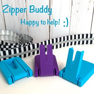 May include: Three plastic zipper pull tools, two in bright blue and one in purple, positioned in front of a coiled black and white striped zipper. The text Zipper Buddy Happy to help! is displayed in blue above the items.