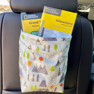 Green multicolor hanging organizer bag with mountains and trees printed. Bag is holding yellow National Geographic maps from Yellowstone and Grand Teton National Park. The fabric is oil cloth. The bag has a strap and is hanging on a black car seat.