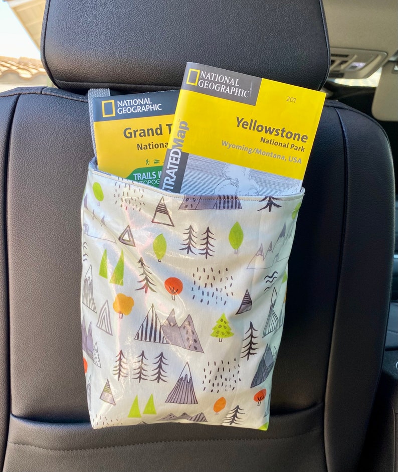 Green multicolor hanging organizer bag with mountains and trees printed. Bag is holding yellow National Geographic maps from Yellowstone and Grand Teton National Park. The fabric is oil cloth. The bag has a strap and is hanging on a black car seat.