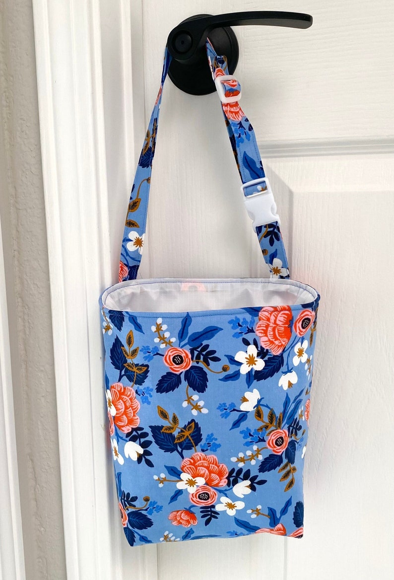 Floral car organizer or litter bag hanging on doorknob of white door. Bag has a strap with white buckle. Fabric is blue with ;ink and white flowers. Interior is waterproof white nylon fabric. Exterior fabric is rifle paper co. Doorknob is black.