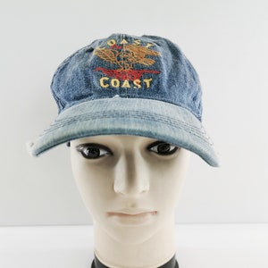 May include: A blue denim baseball cap featuring an embroidered design of a golden eagle above the word COAST in yellow thread and a red line underneath, with the word COAST repeated in yellow thread below the red line on the front panel.