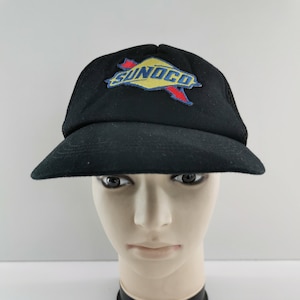 Puede incluir: Gorra de camionero negra con el logotipo de Sunoco bordado en la parte delantera. El logotipo presenta la palabra "SUNOCO" en letras azules con un fondo amarillo y detalles en rojo. La gorra tiene una visera curva y una parte trasera de malla.
