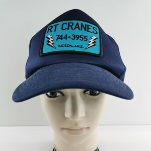 Könnte beinhalten: Eine marineblaue Trucker-Cap mit gebogenem Schirm. Die Kappe hat einen rechteckigen Aufnäher mit dem Text "RT CRANES" über der Telefonnummer "744-3955" und der Stadt "TUCSON, ARIZ." Der Aufnäher hat einen hellblauen Hintergrund mit zwei Blitzgrafiken.