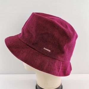 K&ouml;nnte beinhalten: Ein bordeauxfarbener Bucket Hat aus strukturiertem Stoff. Der Hut hat eine kurze Krempe und das Wort "KANGOL" in wei&szlig;en Buchstaben. Der Hut wird auf einem Schaufensterkopf pr&auml;sentiert.