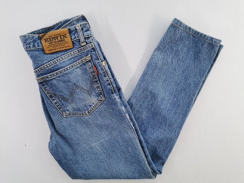 Puede incluir: Vaqueros de mezclilla azul claro con un dise&ntilde;o cl&aacute;sico de cinco bolsillos. Cuenta con un parche de cuero marr&oacute;n con el logotipo "Edwin US Classic" y una etiqueta roja en el bolsillo trasero. Los vaqueros son de mezclilla duradera.
