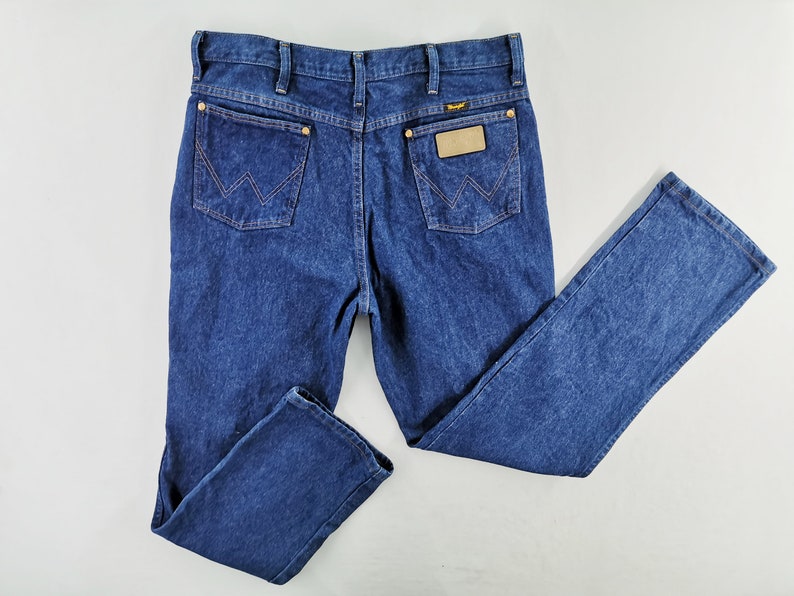 Puede incluir: Vaqueros de mezclilla azul oscuro con un dise&ntilde;o cl&aacute;sico de cinco bolsillos. Presentan el logotipo de Wrangler en el bolsillo trasero y un parche de cuero. Los vaqueros tienen un corte de pierna recta y est&aacute;n hechos de un material de mezclilla duradero.