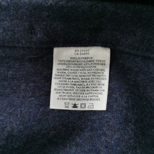 K&ouml;nnte beinhalten: Nahaufnahme eines wei&szlig;en Kleidungsetiketts mit schwarzem Text. Das Etikett enth&auml;lt Angaben zur Stoffzusammensetzung: 100 % Schurwolle, 65 % Polyester und 35 % Baumwolle. Auch Waschanweisungen sind enthalten. Das Etikett befindet sich auf einem dunkelblauen Stoff.