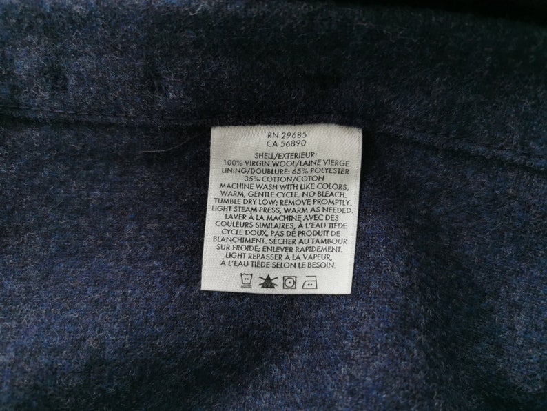 K&ouml;nnte beinhalten: Nahaufnahme eines wei&szlig;en Kleidungsetiketts mit schwarzem Text. Das Etikett enth&auml;lt Angaben zur Stoffzusammensetzung: 100 % Schurwolle, 65 % Polyester und 35 % Baumwolle. Auch Waschanweisungen sind enthalten. Das Etikett befindet sich auf einem dunkelblauen Stoff.