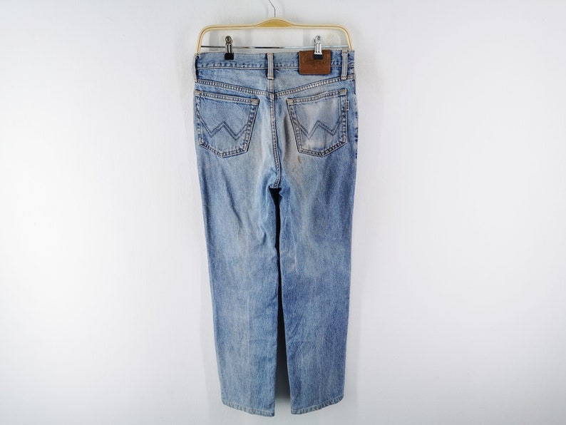 Puede incluir: Vaqueros de mezclilla azul claro con un dise&ntilde;o cl&aacute;sico de cinco bolsillos, colgados de una percha de madera. Los vaqueros presentan un lavado descolorido y un parche de cuero marr&oacute;n en la parte trasera. Los bolsillos traseros tienen una costura en 'W'.