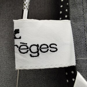 May include: Close-up of a white label with the text "rēges" in black, attached to a dark gray garment. The garment has a textured appearance and is hanging on a silver hanger. The label is rectangular with a stitched border.