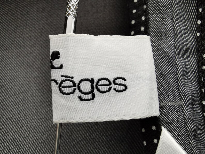 May include: Close-up of a white label with the text "rēges" in black, attached to a dark gray garment. The garment has a textured appearance and is hanging on a silver hanger. The label is rectangular with a stitched border.