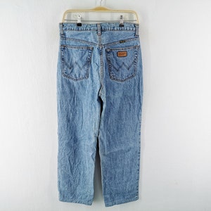 May include: Light blue denim jeans hanging on a hanger. The jeans have a classic five-pocket design, a straight leg, and a brown leather patch with the brand name. The back pockets feature a distinctive stitching pattern.