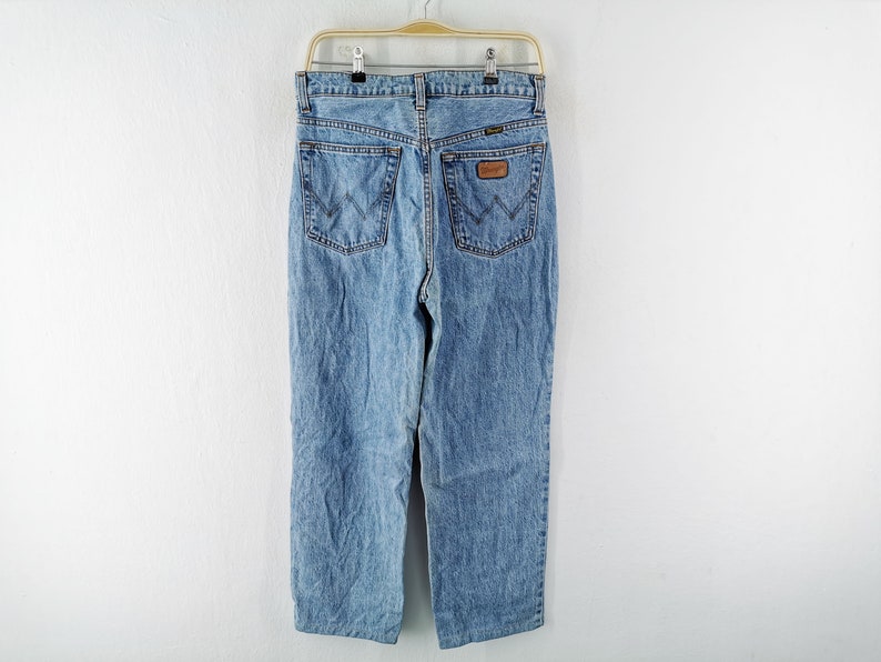 May include: Light blue denim jeans hanging on a hanger. The jeans have a classic five-pocket design, a straight leg, and a brown leather patch with the brand name. The back pockets feature a distinctive stitching pattern.