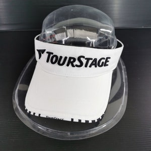 May include: White visor with the text "TOURSTAGE" in black. The visor has a black and white checkered pattern on the brim. The visor is displayed on a clear plastic holder.