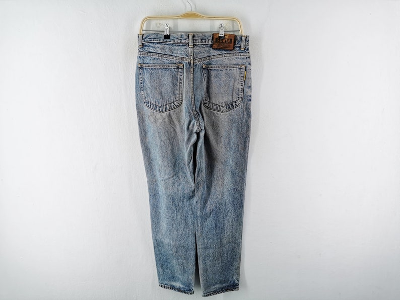 Op de afbeelding: Lichtblauwe denim jeans met een vervaagde wassing, hangend aan een houten hanger. De jeans hebben een klassiek ontwerp met vijf zakken, een leren patch en een rechte pijp. De achterzakken hebben een decoratieve steek.