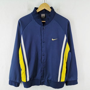 May include: A navy blue button-up jacket with white and yellow side stripes. The jacket has a small yellow Nike logo on the chest. It is hanging on a wooden hanger against a white background.
