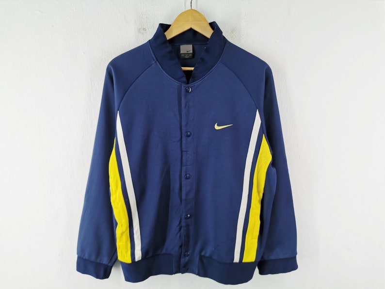 May include: A navy blue button-up jacket with white and yellow side stripes. The jacket has a small yellow Nike logo on the chest. It is hanging on a wooden hanger against a white background.