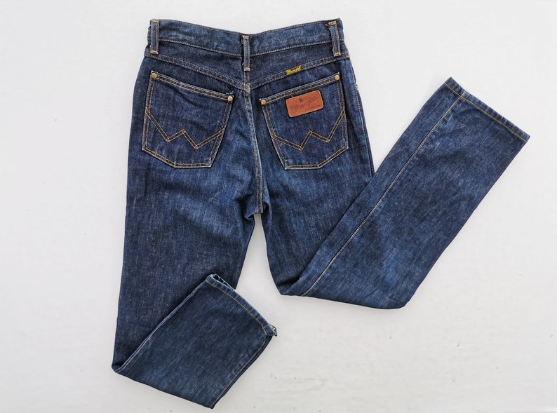 Op de afbeelding: Donkerblauwe denim jeans met een klassiek vijf-zakken ontwerp. Voorzien van een bruine leren patch en het Wrangler-logo. De achterzakken hebben de kenmerkende Wrangler-stiksels.